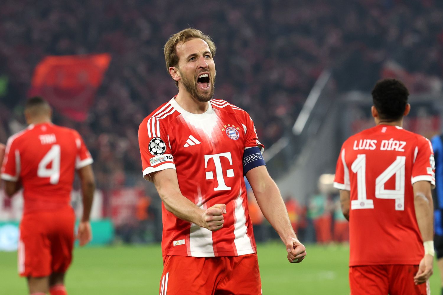 FC Bayern player in red cheers on the pitch during a match, surrounded by teammates and fans, at an important Champions League match.